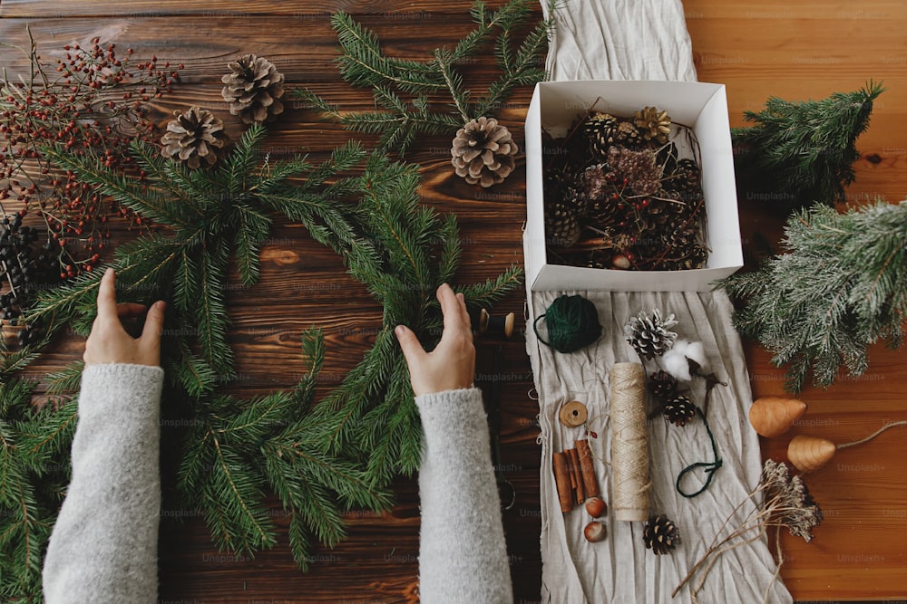 소박한 크리스마스 화환을 평평하게 만듭니다. 소나무 콘, 딸기, 자연 축제 장식이 있는 소박한 나무 테이블에 녹색 전나무 가지를 들고 있는 손. 계절 겨울 워크샵, 휴일 강림