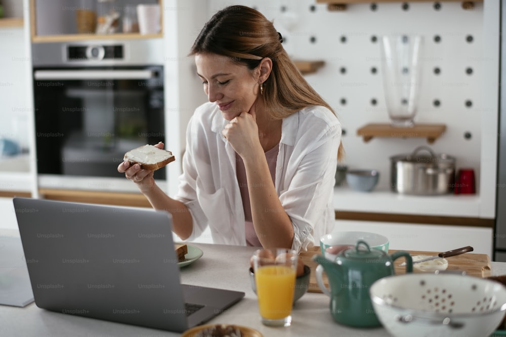 Young woman in kitchen. Beautiful woman having video call while eating the sandwich.