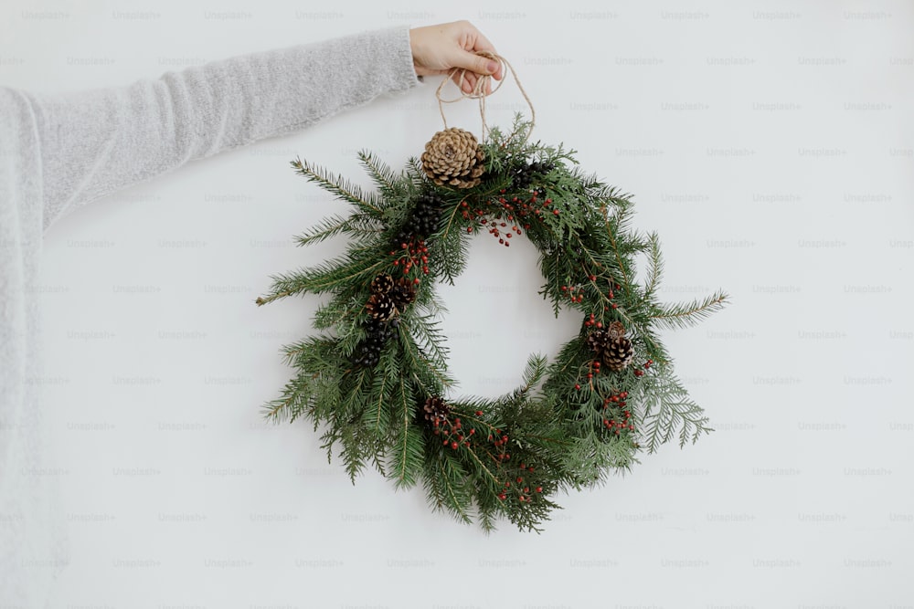 Rustic christmas wreath in female hands on background of white wall. Merry Christmas and Happy Holidays! Woman in stylish  sweater holding modern christmas wreath, holiday advent
