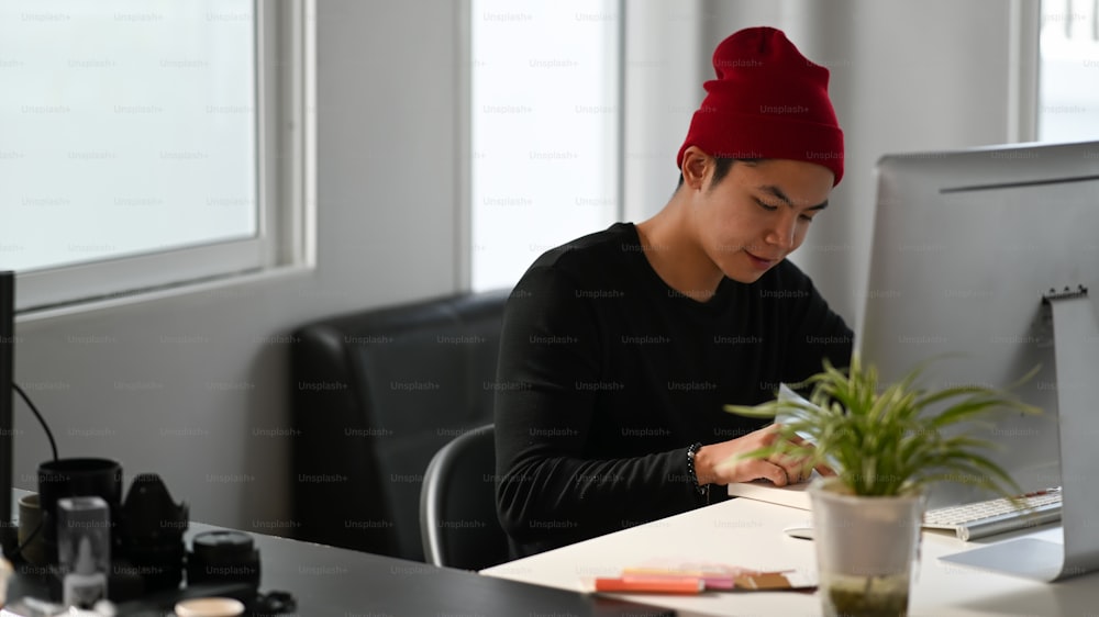 Un graphic designer creativo maschio sta leggendo un libro mentre è seduto davanti al computer sul posto di lavoro.