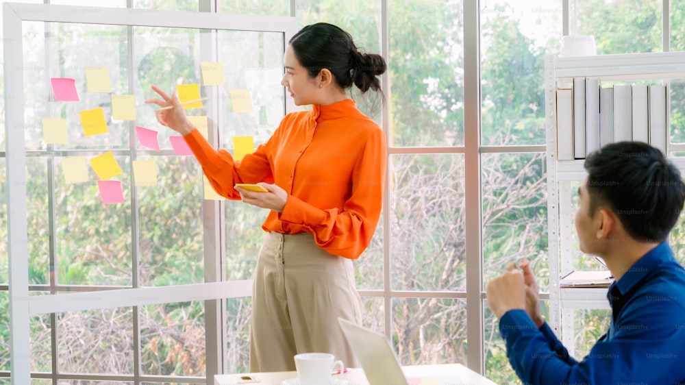 Business people work on project planning board in office and having conversation with coworker friend to analyze project development . They use sticky notes posted on glass wall to make it organized .