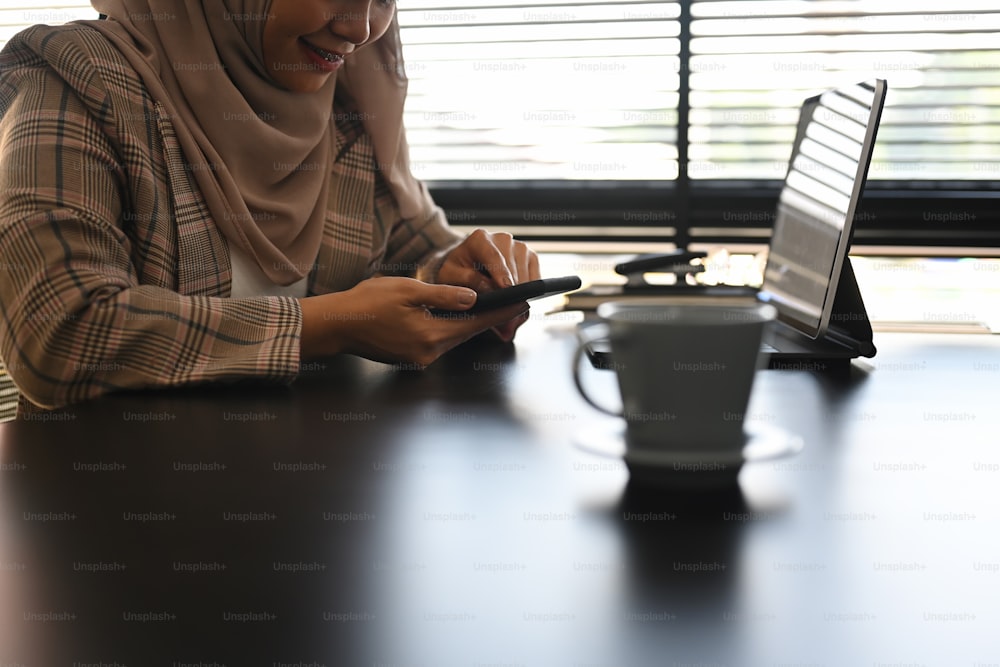 Ausschnittaufnahme einer schönen asiatischen muslimischen Geschäftsfrau benutzt ein Smartphone, während sie an ihrem Arbeitsplatz sitzt.