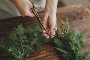 Making rustic christmas wreath, seasonal winter workshop. Florist hands holding scissors and berries, making christmas wreath on wooden table with natural festive decorations. Holiday advent