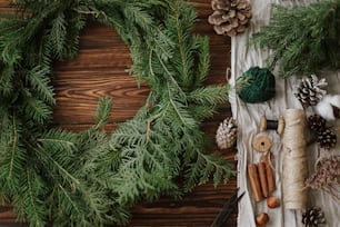 Coroa de Natal com bagas, ramos verdes, pinhas e decorações festivas naturais, tesoura, barbante. Fazendo grinalda de Natal rústica na mesa de madeira flat lay, feriado sazonal adven