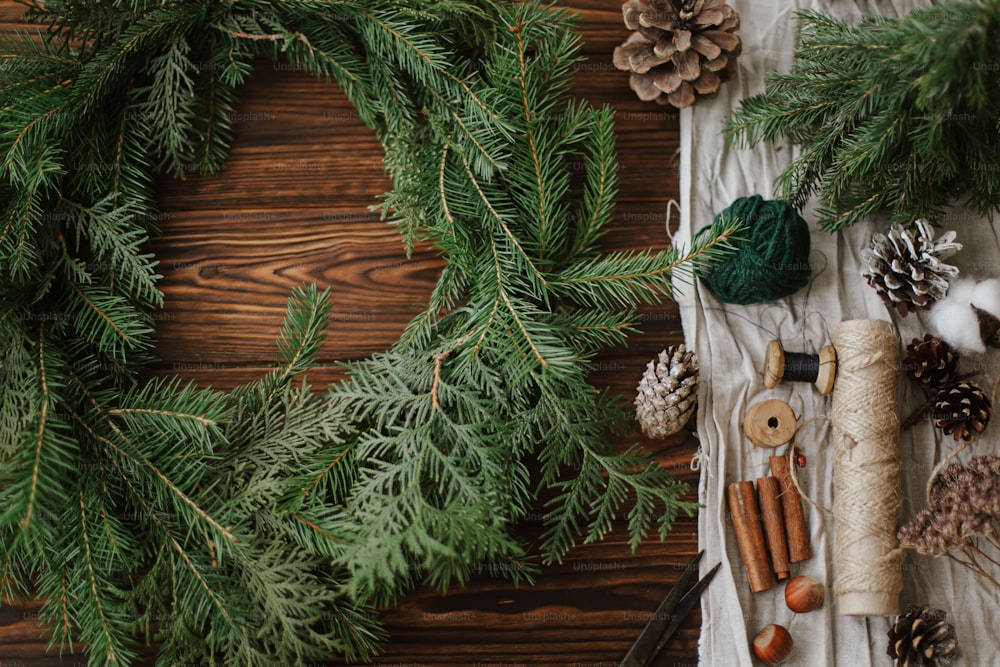 Corona navideña con bayas, ramas verdes, piñas y decoraciones festivas naturales, tijeras, cordel. Hacer una corona rústica de Navidad en una mesa de madera plana, advenido de vacaciones de temporada