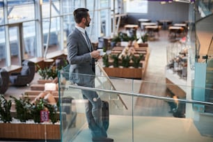 Foto in voller Länge eines männlichen Geschäftsmanns, der eine Tasse Kaffee trinkt, während er auf die Flughafenlounge starrt