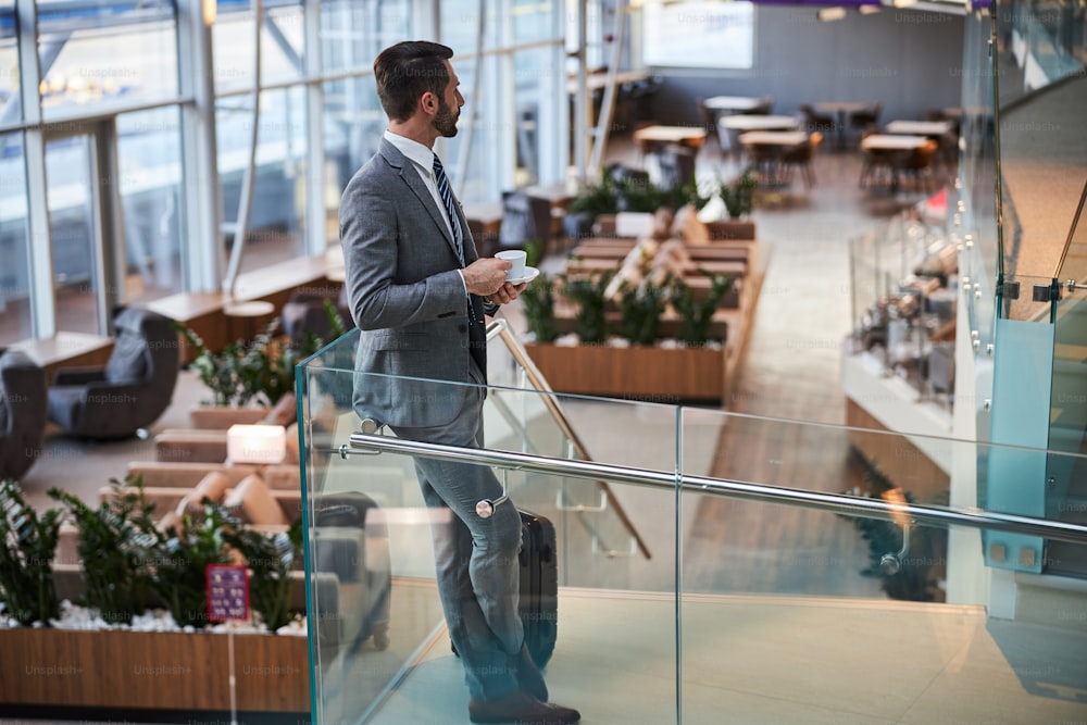 Full-length photo of a male businessperson drinking a cup of coffee while staring at the airport lounge