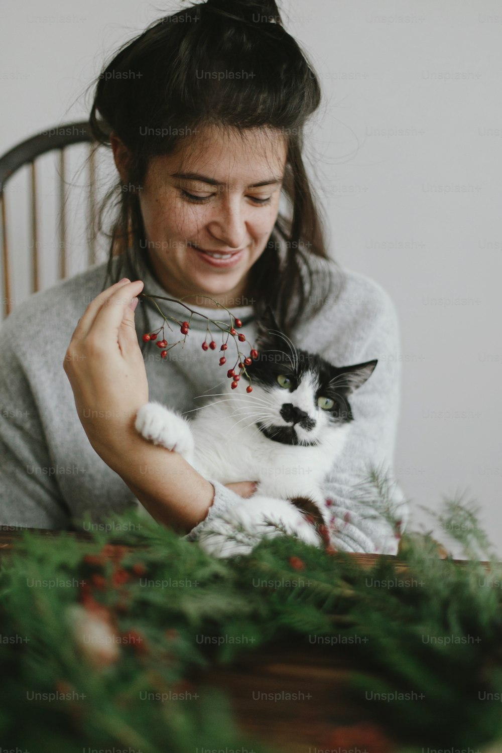 Faire une couronne de Noël avec un chat mignon à la maison, l’avent des vacances. Femme heureuse faisant une couronne de Noël avec une adorable aide féline, tenant des baies rouges et des branches vertes. Image authentique et élégante