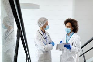African American female doctor and her colleagues talking while analyzing medical report and wearing protective face mask in a hallway at the clinic.