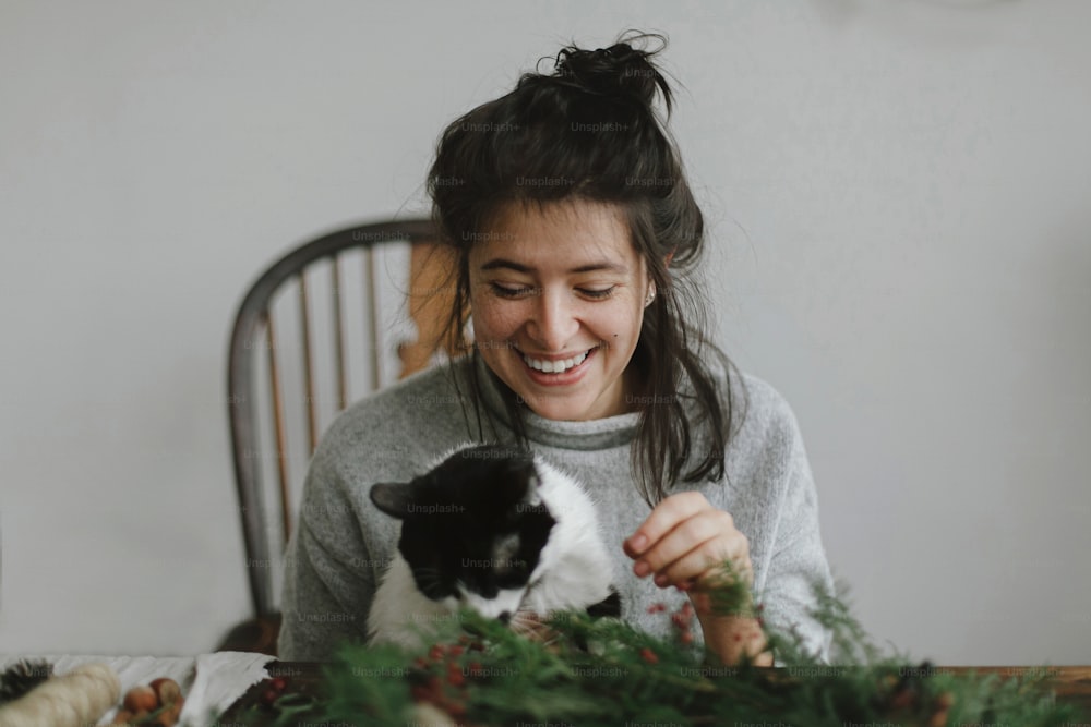 かわいい猫は、赤い果実と緑の枝、休日のアドベントを保持し、素朴なクリスマスリースを作る若い幸せな女性を助けます。スタイリッシュで本格的なイメージ、ペットと自宅での休日。楽しい休暇をお過ごしください
