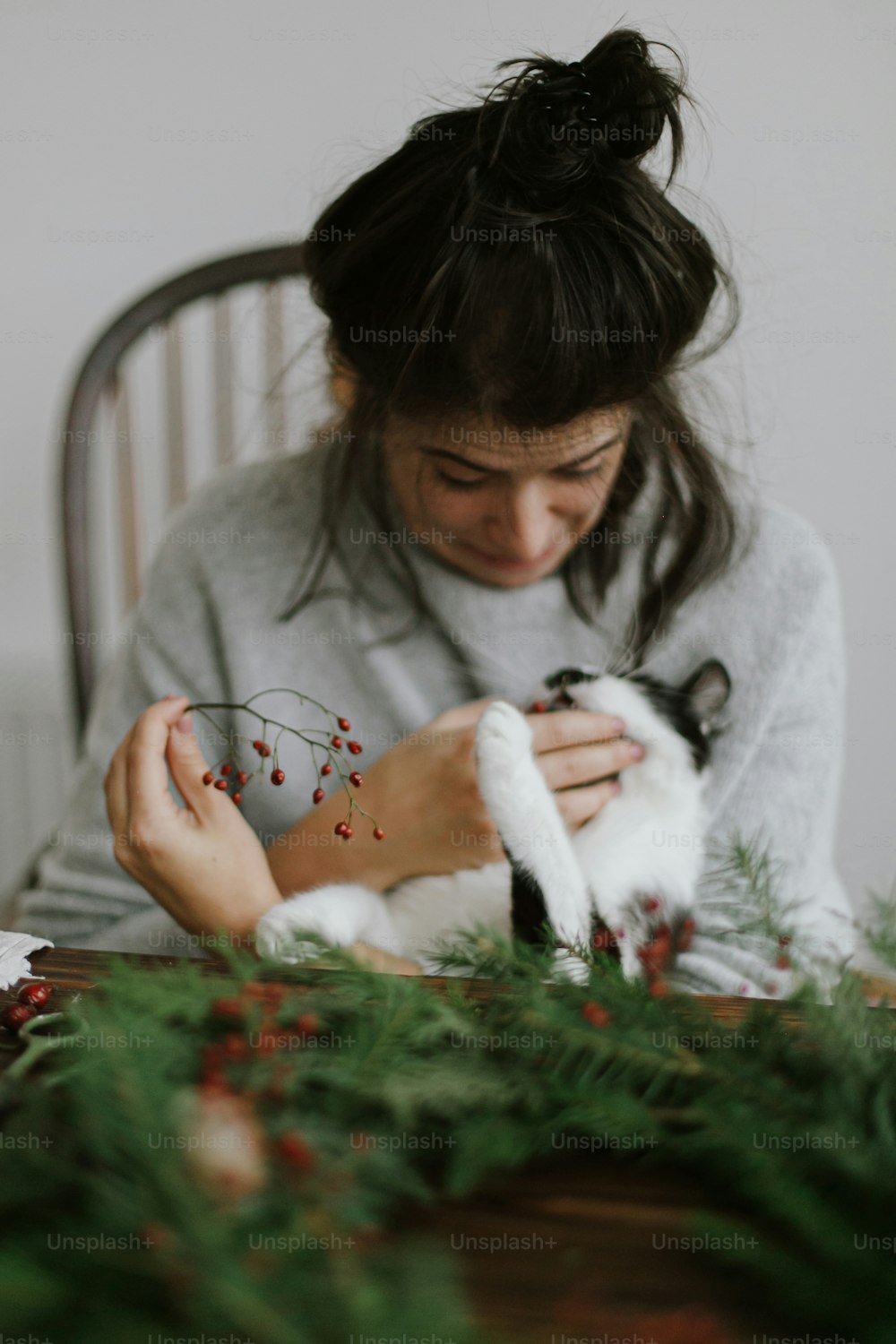 빨간 열매와 녹색 가지를 들고 소박한 크리스마스 화환을 만드는 젊은 행복한 여자를 돕는 귀여운 고양이. 재미있는 정통 가정의 순간, 애완 동물 및 휴일. 즐거운 휴일 보내세요