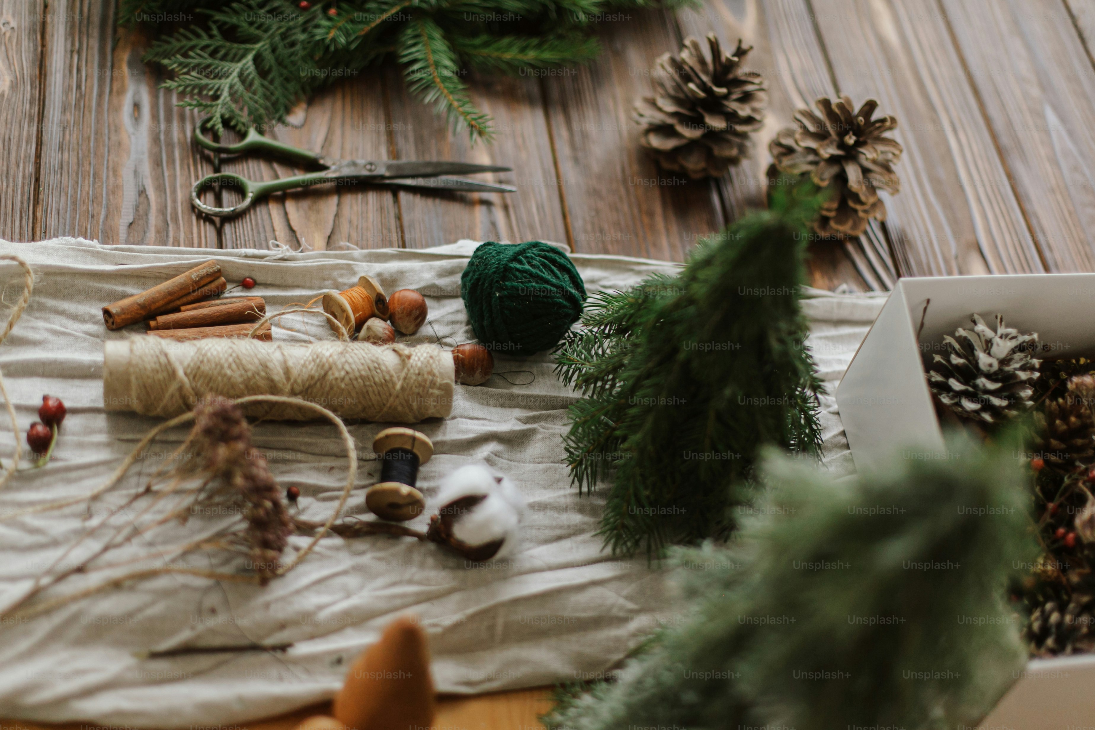 Craft supplies including pine cones, cinnamon sticks, twine, and greenery on linen
