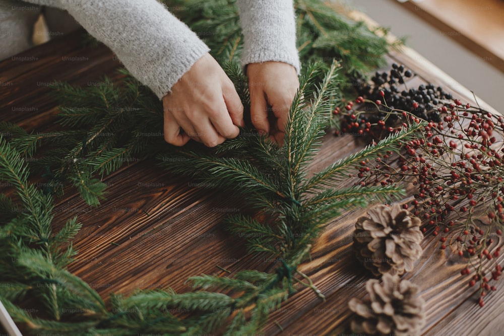 Hands holding green fir branches on rustic wooden table with pine cones, berries, natural festive decorations. Making rustic christmas wreath. Seasonal winter workshop, holiday advent