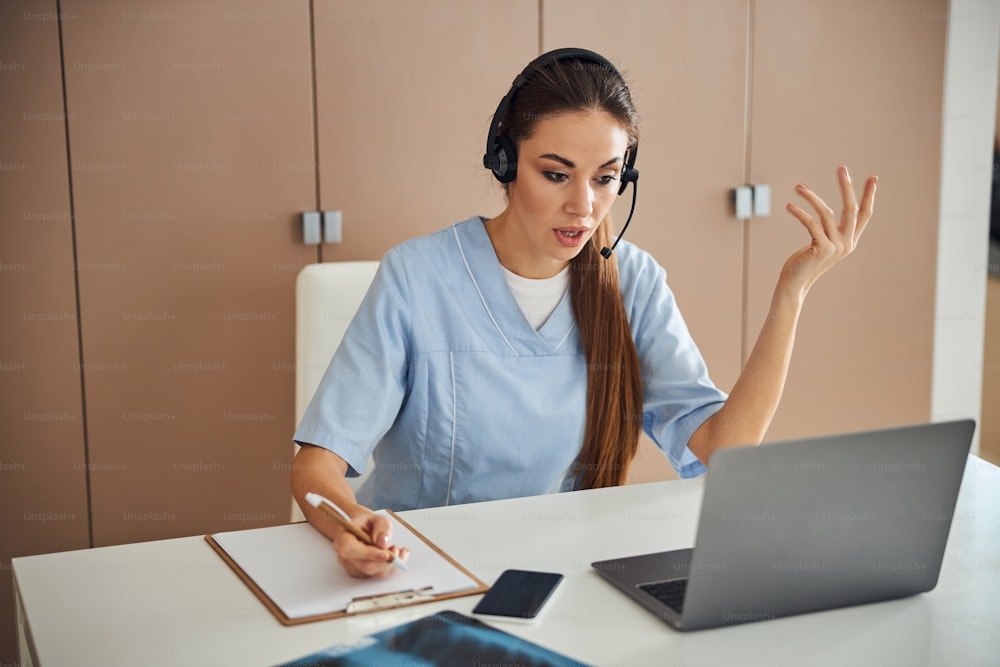 Experienced female doctor wearing scrubs and a headset while talking on a video-call and making gestures