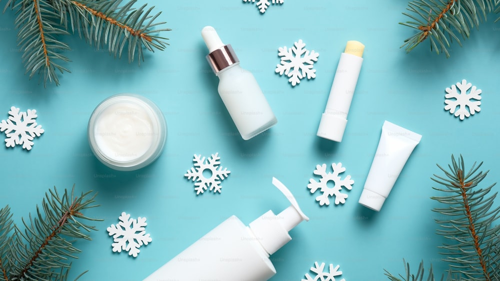 Set of winter skincare cosmetics on blue background. Packaging design template. Moisturizer cream jar, lipstick, pump bottle, tube, snowflakes and fir tree branches. Flat lay, top view.