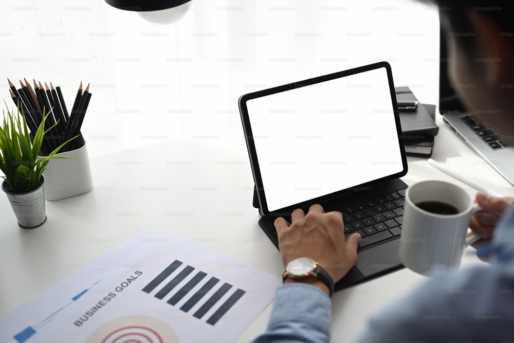 Abgeschnittene Aufnahme eines Geschäftsmannes, der Kaffee trinkt und am Tablet-Computer mit digitalem Layer-Graph-Informationsdiagramm auf dem Bürotisch arbeitet.