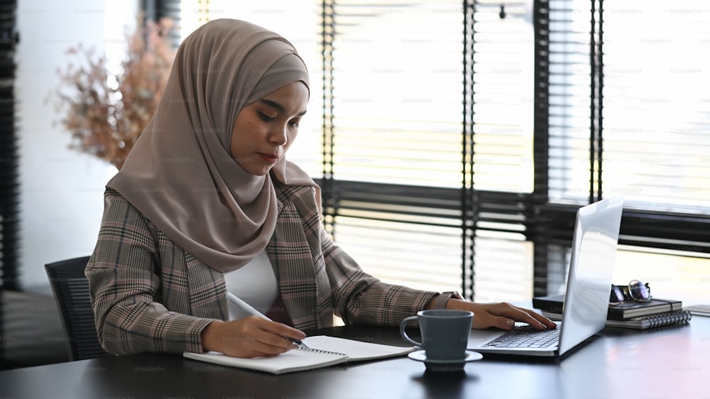 ヒジャブをかぶったイスラム教徒のビジネスウーマンが、現代のオフィスでスタートアッププロジェクトにラップトップで取り組んでいます。