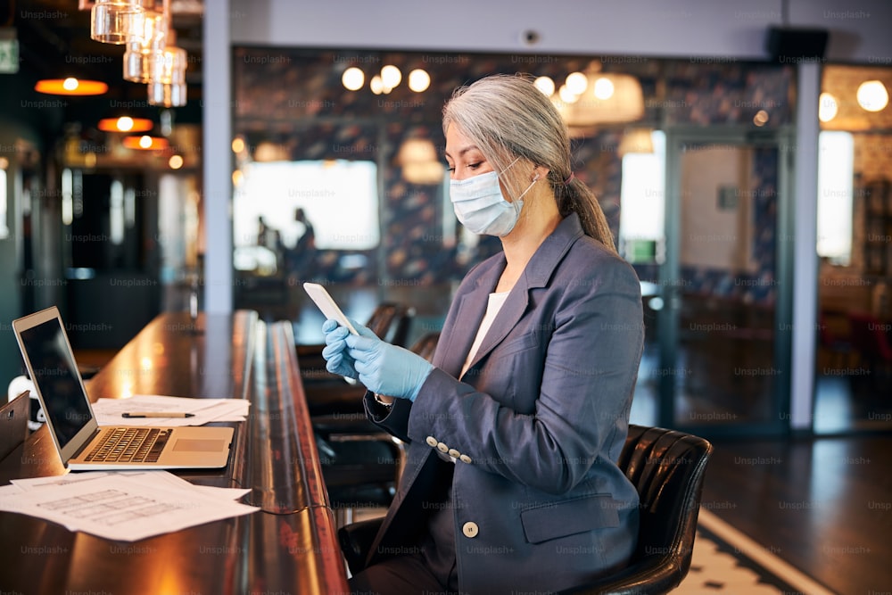 Elegant woman in sterile gloves texting message on smartphone while sitting at the counter with laptop and papers