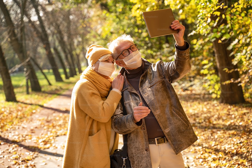 Smiling senior couple using digital tablet in autumn park. Senior couple wearing protective medical mask for protection from virus. Health, quarantine and pandemic concept.