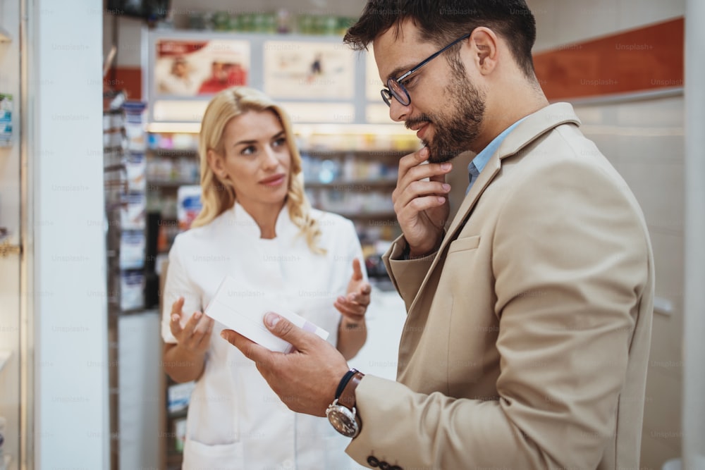 Young business man choosing and buying drugs in a drugstore while talking with attractive female pharmacist. She helping him with expert advices.