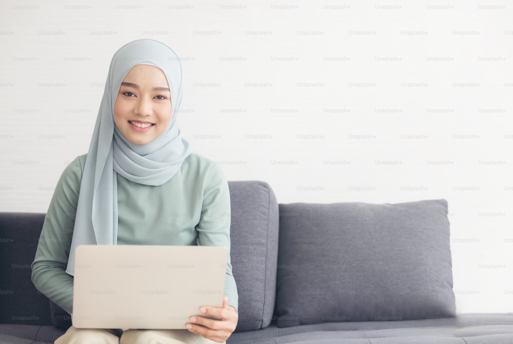 Portrait of a young happy muslim woman in hijab working at home using laptop computer while sitting on sofa.