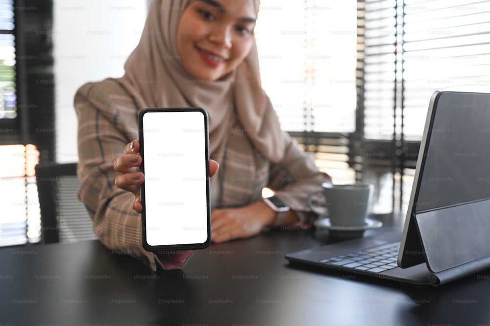 Foto cortada da bela empresária muçulmana asiática está mostrando um smartphone com tela isolada sobre a mesa do escritório.