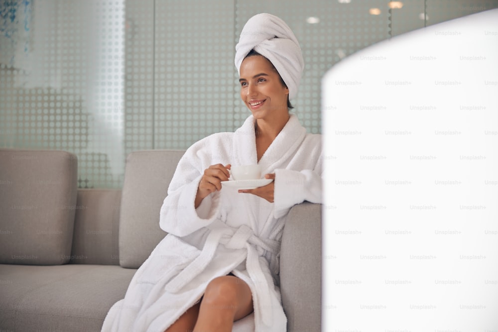 Happy pretty lady in white robe sitting on sofa and holding hot drink while relaxing in spa salon. Copy space