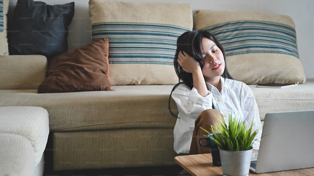 Una mujer joven siente un fuerte dolor de cabeza o está agotada por el exceso de trabajo mientras está sentada en la sala de estar.