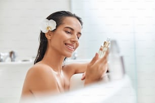 Charming lady with white flower in her hair looking at skincare product and smiling while relaxing in bathtub