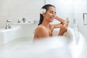 Jolie dame avec une orchidée blanche dans les cheveux regardant un produit de soin de la peau et souriant tout en se relaxant dans la baignoire
