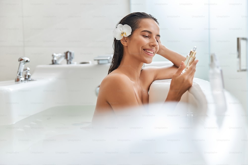 Attractive lady with white orchid in her hair looking at skincare product and smiling while relaxing in bathtub