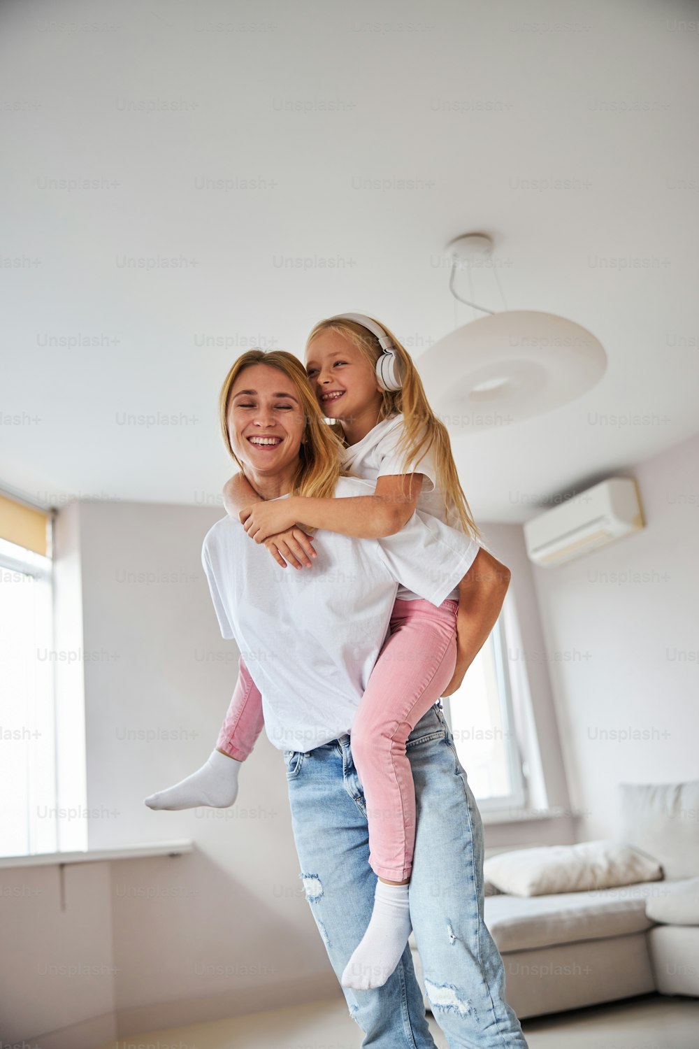 흰 셔츠와 청바지를 입은 행복한 쾌활한 젊은 엄마가 주말에 집에서 시간을 보내는 동안 어린 딸을 등에 안고 있다