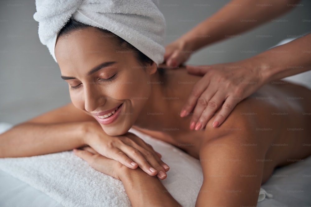 Attractive lady with bath towel on her head lying on massage table and smiling while receiving back massage
