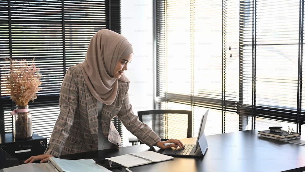 Eine junge arabische Unternehmerin, die einen Hijab trägt, arbeitet online mit einem Laptop in einem modernen Büro.