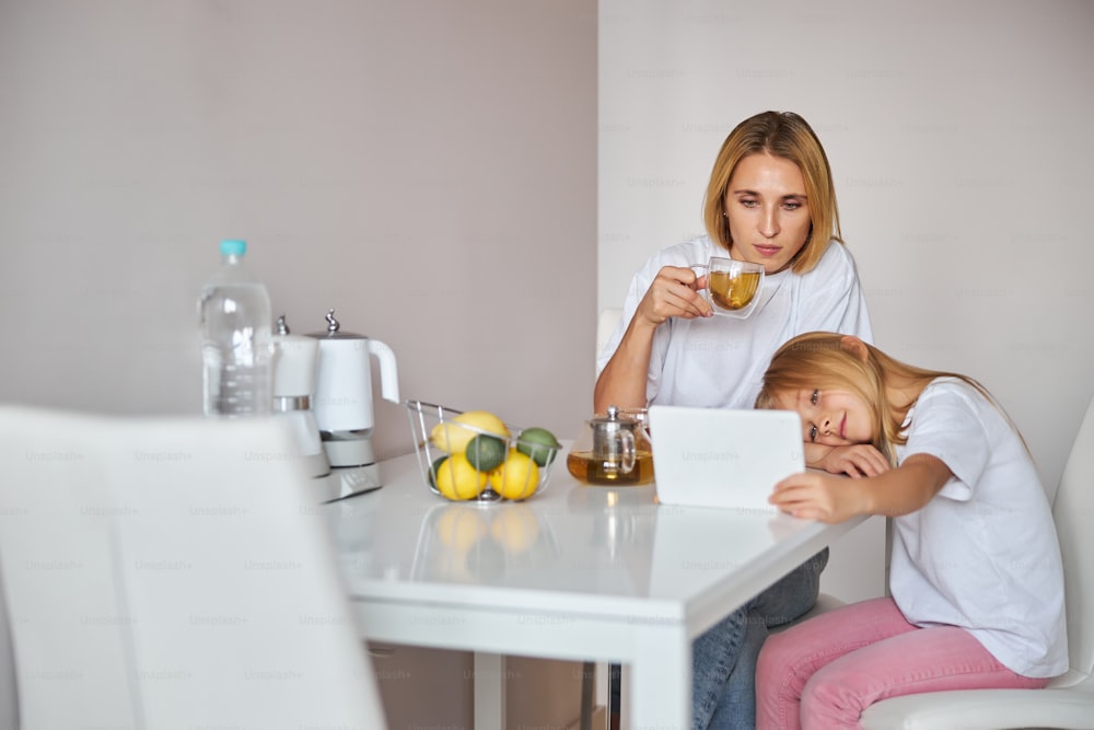 小さな娘が自宅の台所で現代のタブレットで何かを実演している間、飲み物を手にマグカップを持っているスタイリッシュでエレガントな母親の腰の肖像画