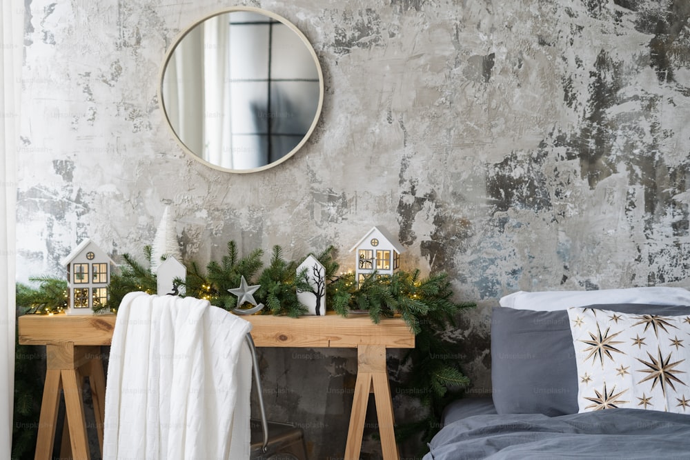 ロフトのインテリア、柔らかく快適なベッド、素朴な壁の近くの木製のテーブルの上の冬の季節の装飾の鏡を備えたモダンなデザインのアパートの寝室の要素