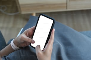 Vista de cerca de las manos de una mujer independiente usando una maqueta de teléfono inteligente mientras está sentada en el sofá. Teléfono móvil con pantalla en blanco para montaje gráfico.