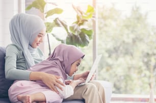 Asian muslim mother and daughter smiling when using laptop together, browsing internet or watching streaming movie
