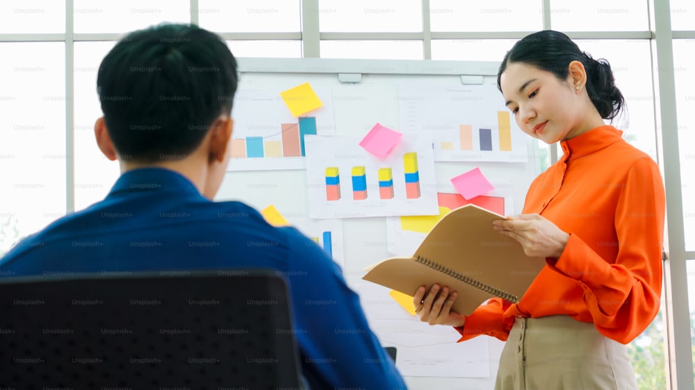 Junge Frau erklärt Geschäftsdaten auf Whiteboard in zwanglosem Büroraum. Die selbstbewusste asiatische Geschäftsfrau berichtet über den Informationsfortschritt eines Geschäftsprojekts an Partner, um die Marktstrategie festzulegen.