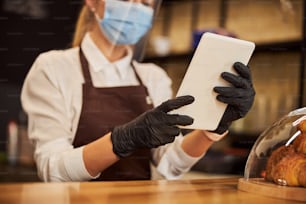 Cropped photo of a woman in protective shield and rubber gloves standing at the bar counter with a white tablet