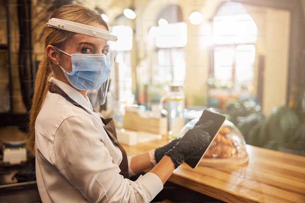 Bonita camarera con escudo protector sobre la máscara médica tocando la pantalla de una tableta en sus manos