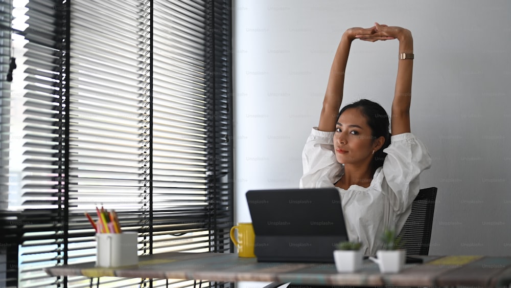A calm smiling businesswoman is relaxing at comfortable office chair and stretching raising hands up.