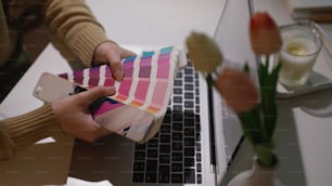 Abgeschnittene Aufnahme von Designerhänden, die Farbmuster halten, während sie mit dem Laptop im Heimbüro arbeiten