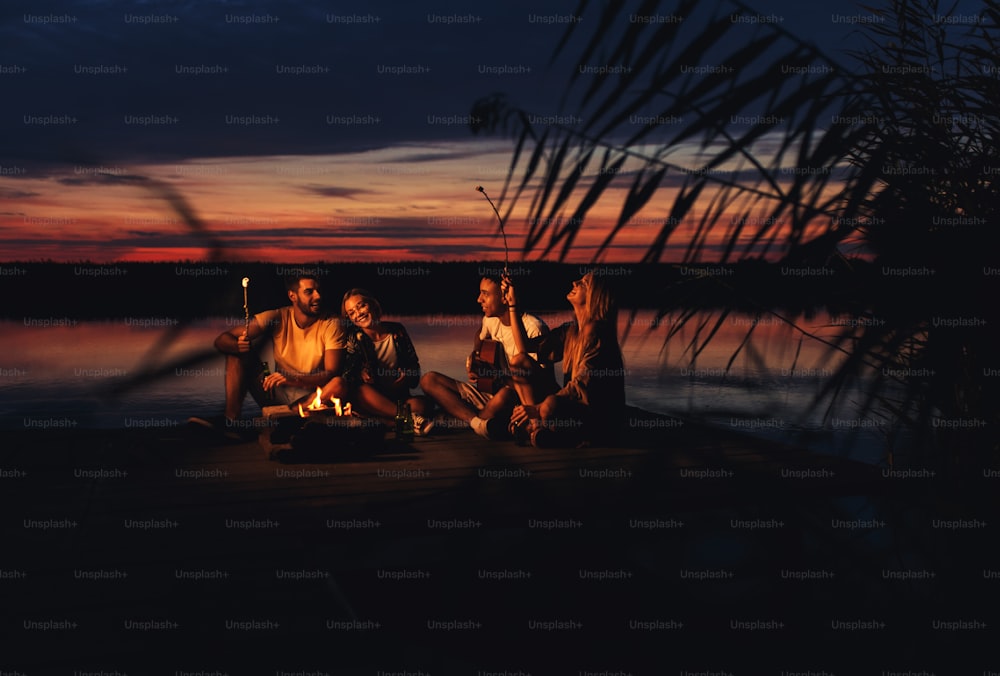 Gruppe junger Freunde, die nachts am See genießen. Sie sitzen um das Feuer herum singen und haben Spaß.