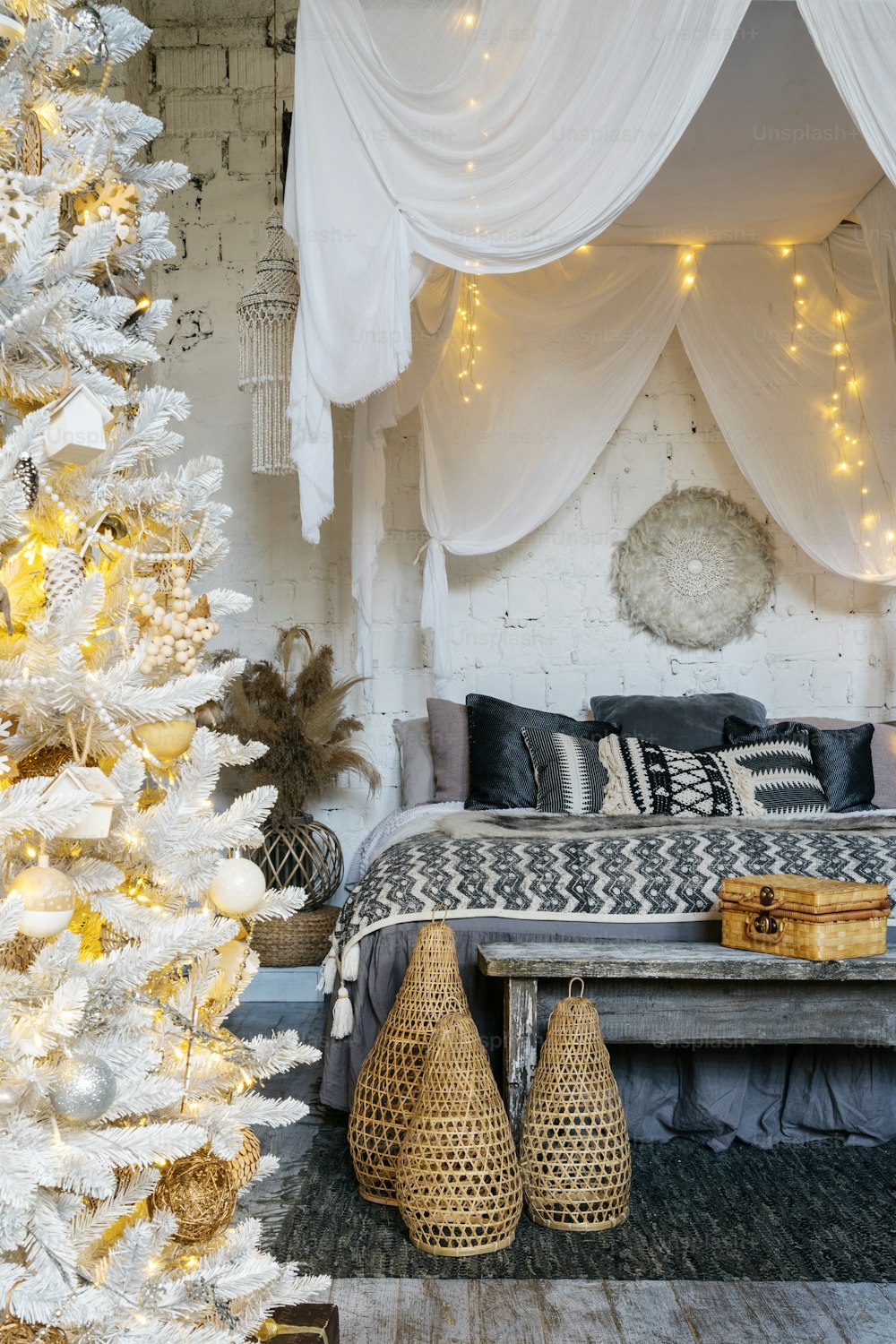 Foto vertical de un dormitorio acogedor en un interior de estilo boho con cama cómoda, cuadros, almohadas y manta, asiento de banco de madera y decoración del hogar cerca del árbol de Navidad decorado