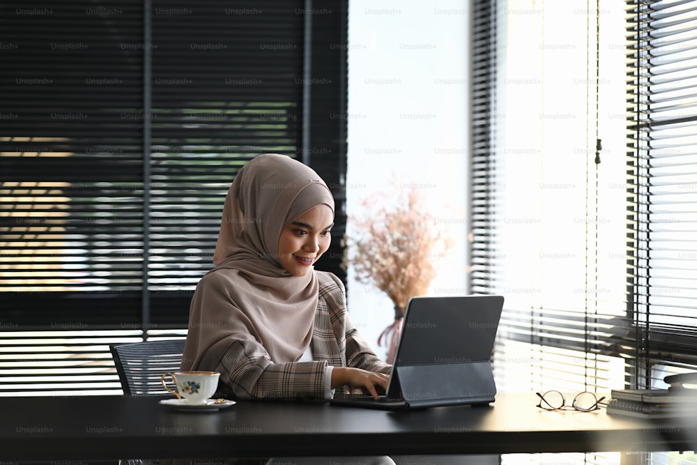 Eine muslimische Frau mit Kopftuch sitzt an ihrem Arbeitsplatz und arbeitet am Laptop in einem modernen Büro.