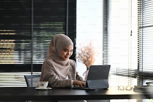 A muslim woman in headscarf is sitting at her workspace and working on laptop in modern office.