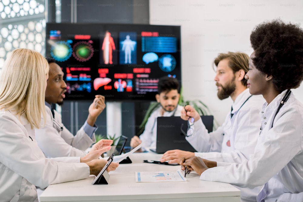 Multiethnic team of five doctors brainstorming or discussing patients records, on the meeting at light office. Focus on young African female doctor, listening her colleagues. Coronavirus pandemy.