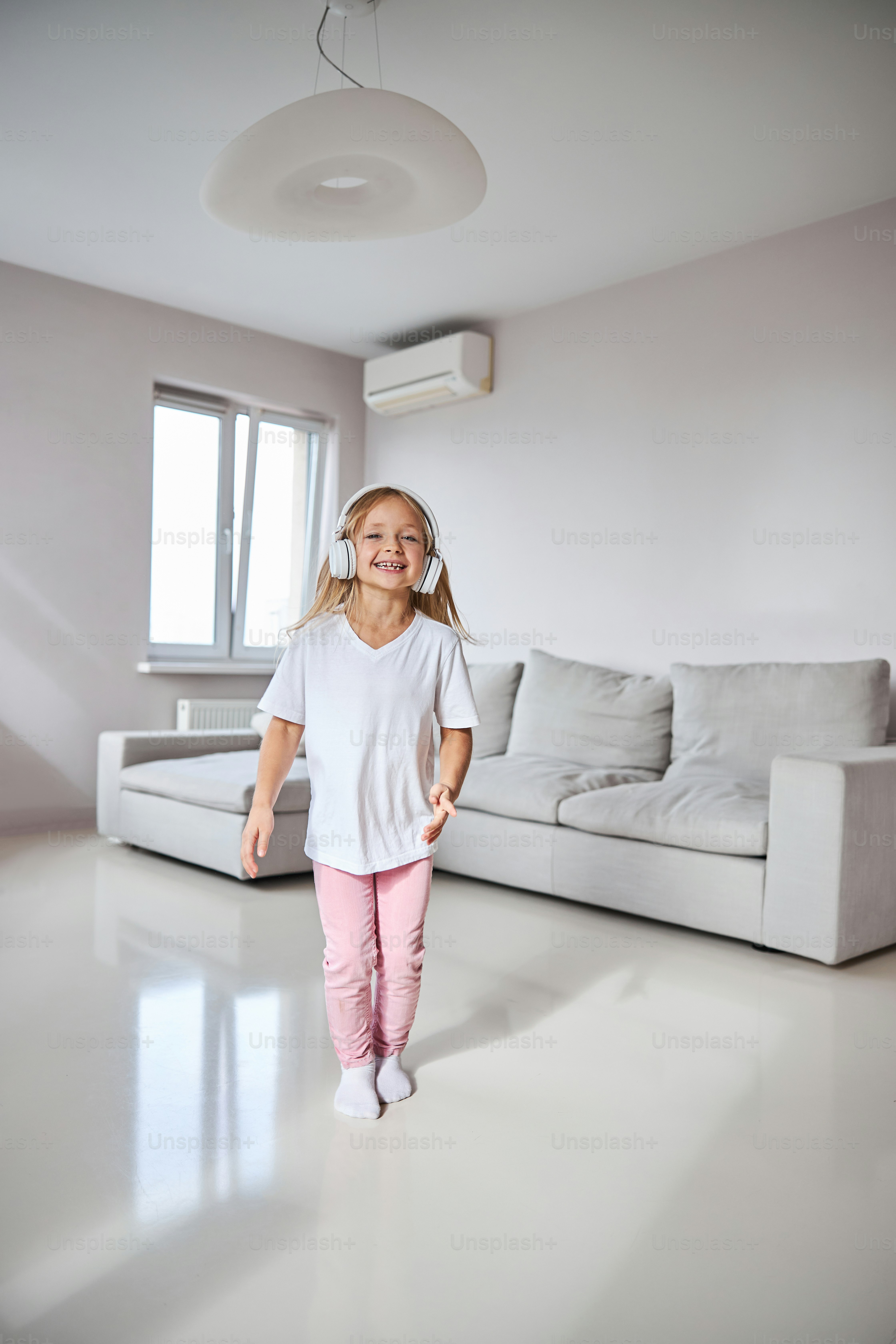 Portrait en pied d’une petite fille souriante heureuse dans des vêtements élégants et des écouteurs dansant dans le salon de la maison