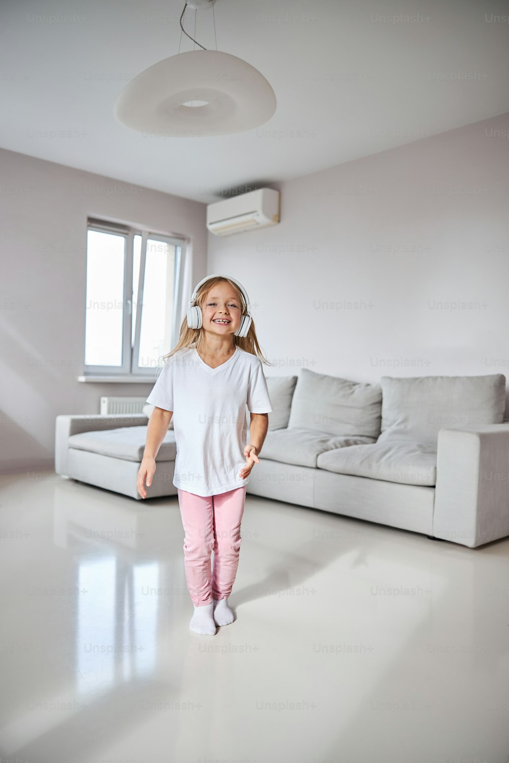 Full length portrait of happy smiling little female child in stylish clothes and headphones dancing in the living room in home